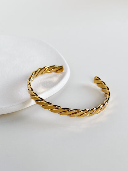 Counter-angled view of the Mila 18k gold PVD twisted cuff bracelet perched gracefully over the curved rim of a minimalist white ceramic tray, showcasing the polished open ends