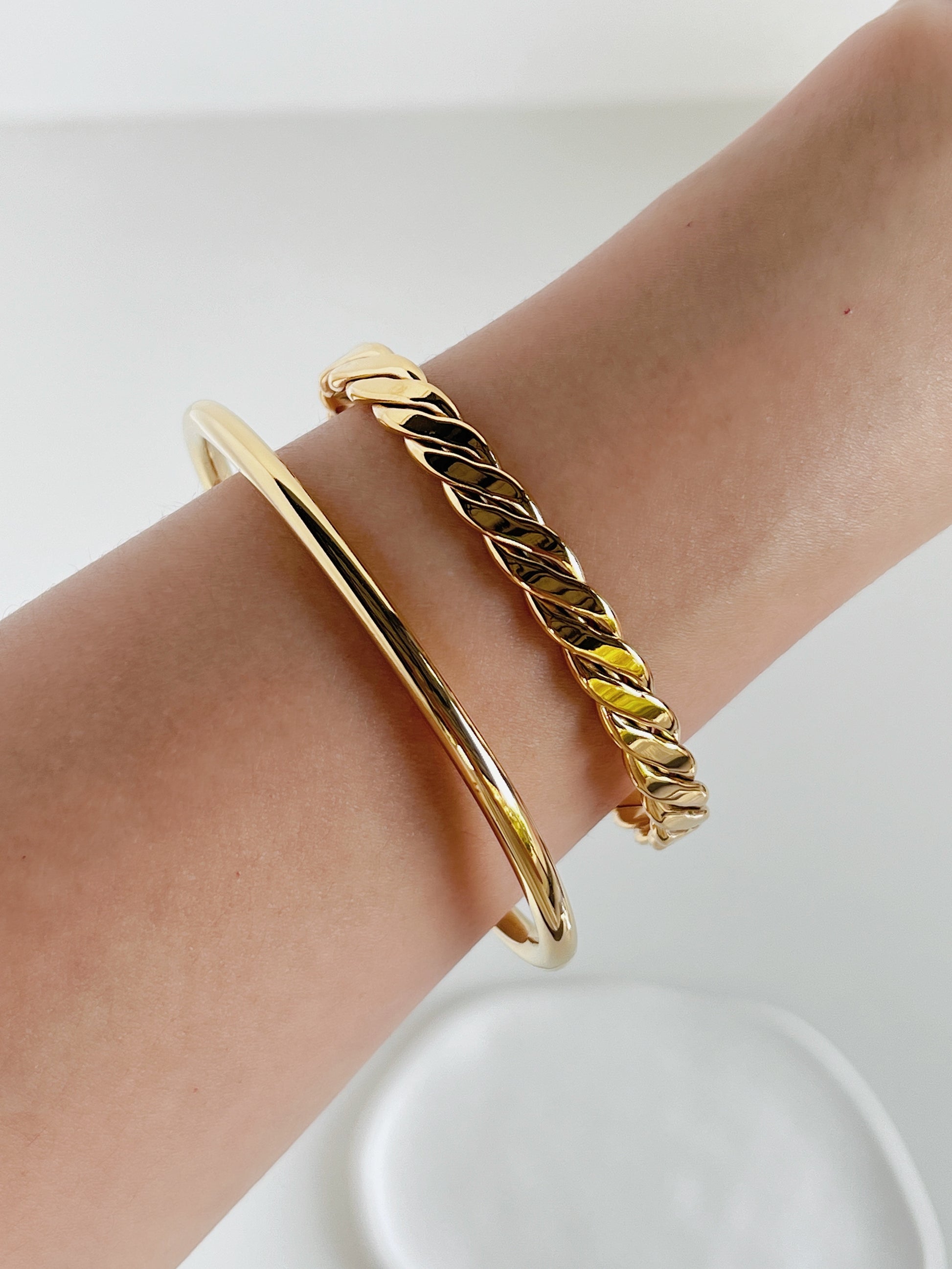 An editorial close-up of a model's wrist showcasing a luxury curation stack featuring the Mila gold braided cuff bangle paired alongside a smooth, solid high-shine gold minimalist cuff bracelet