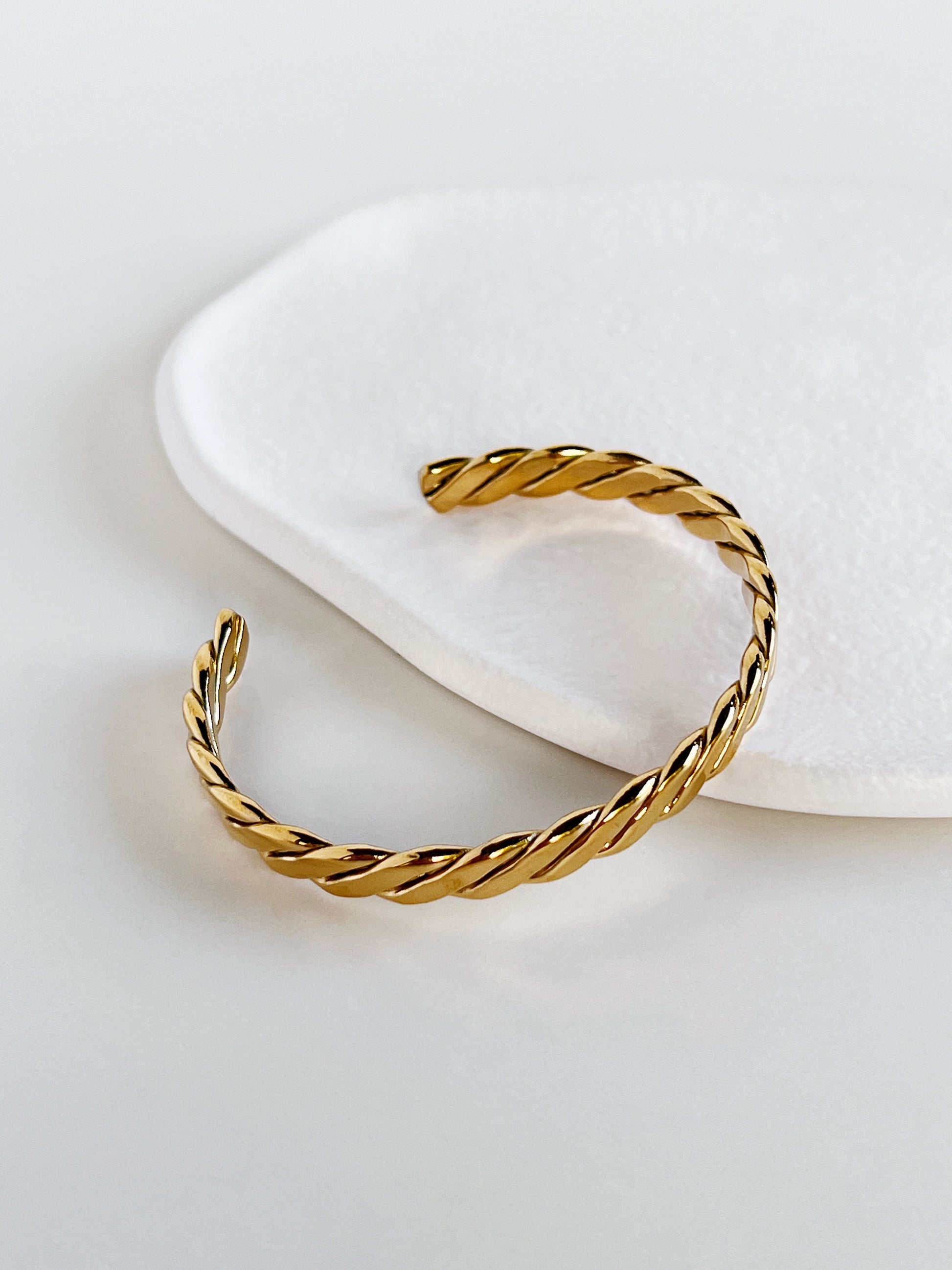 Three-quarter angled product view of the Mila gold braided cuff bangle resting on a textured white porcelain display dish, emphasizing the fluid helical grooves