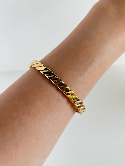 A high-angle, close-up shot of the Mila gold sculpted helical braid cuff bangle worn solo on a model's bare arm, showcasing how the continuous twisted ridges reflect crisp natural light