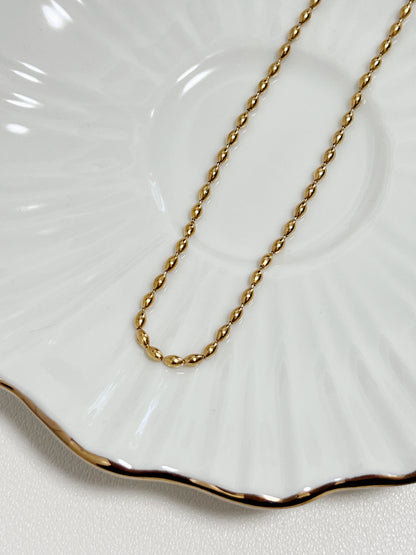 Macro view focusing on the mirror polished finish of the gold rice bead chain resting on a white ceramic surface