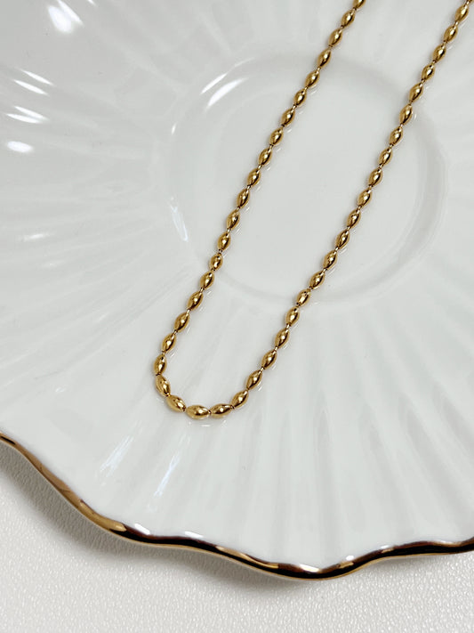 Macro view focusing on the mirror polished finish of the gold rice bead chain resting on a white ceramic surface