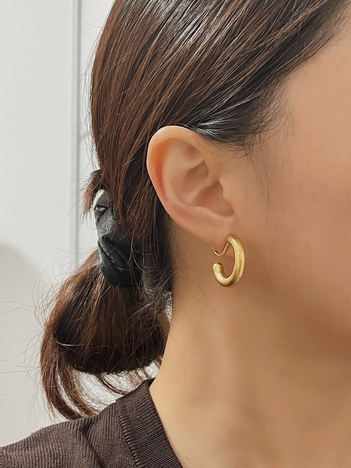 Side profile of a model wearing the Zora chunky brushed gold clip-on hoop earrings, styled with an undone messy low bun and an espresso brown knit top.