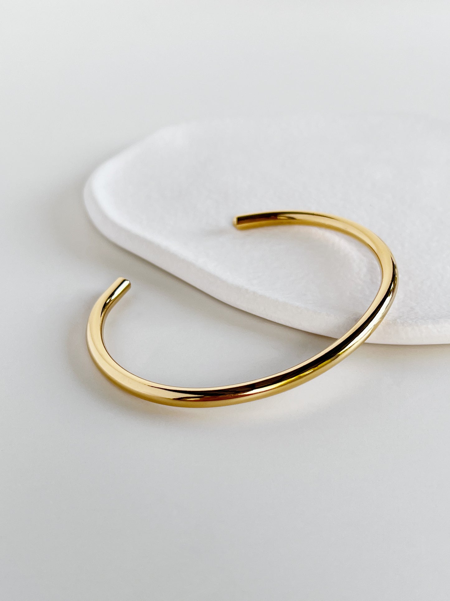 Close-up product view of the polished gold minimal cuff bangle placed diagonally on a stone surface, accentuating the luminous 18K gold PVD coating.