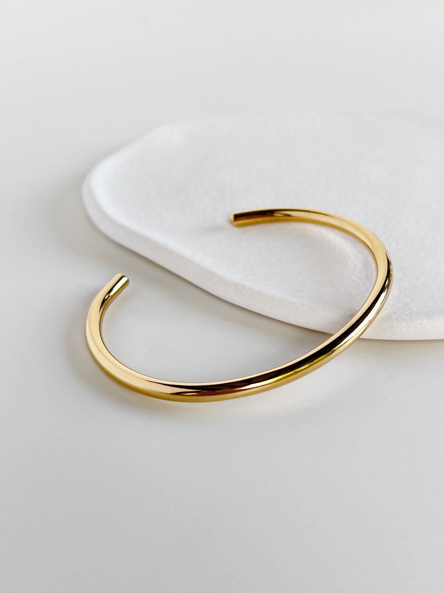 Close-up product view of the polished gold minimal cuff bangle placed diagonally on a stone surface, accentuating the luminous 18K gold PVD coating.