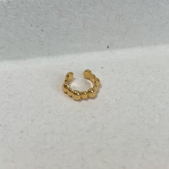 A gold beaded ear cuff displayed on a textured white surface.