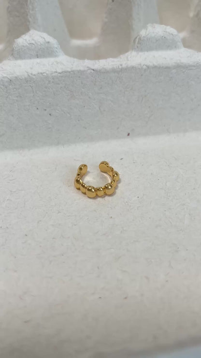 A gold beaded ear cuff displayed on a textured white surface.