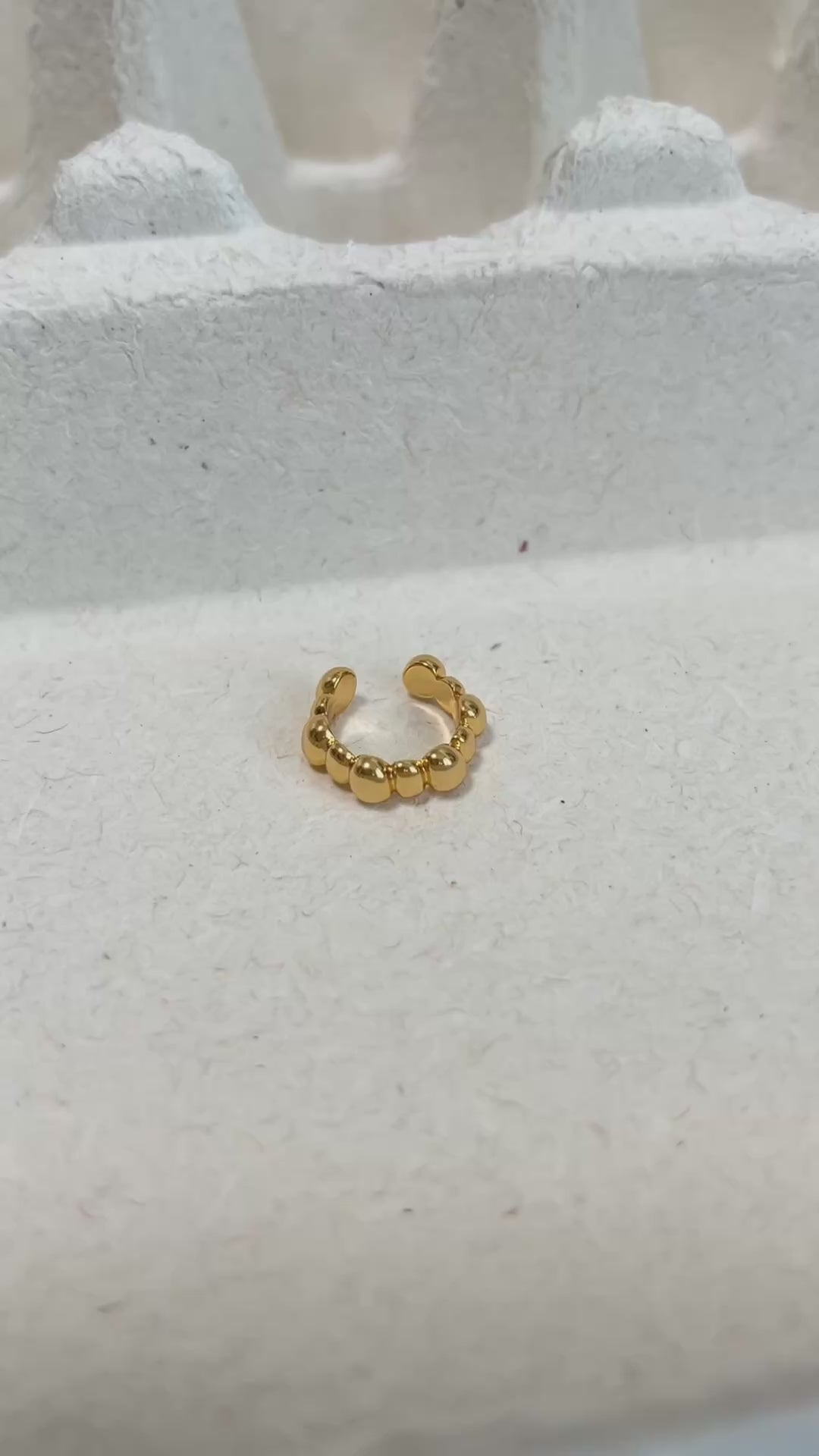 A gold beaded ear cuff displayed on a textured white surface.