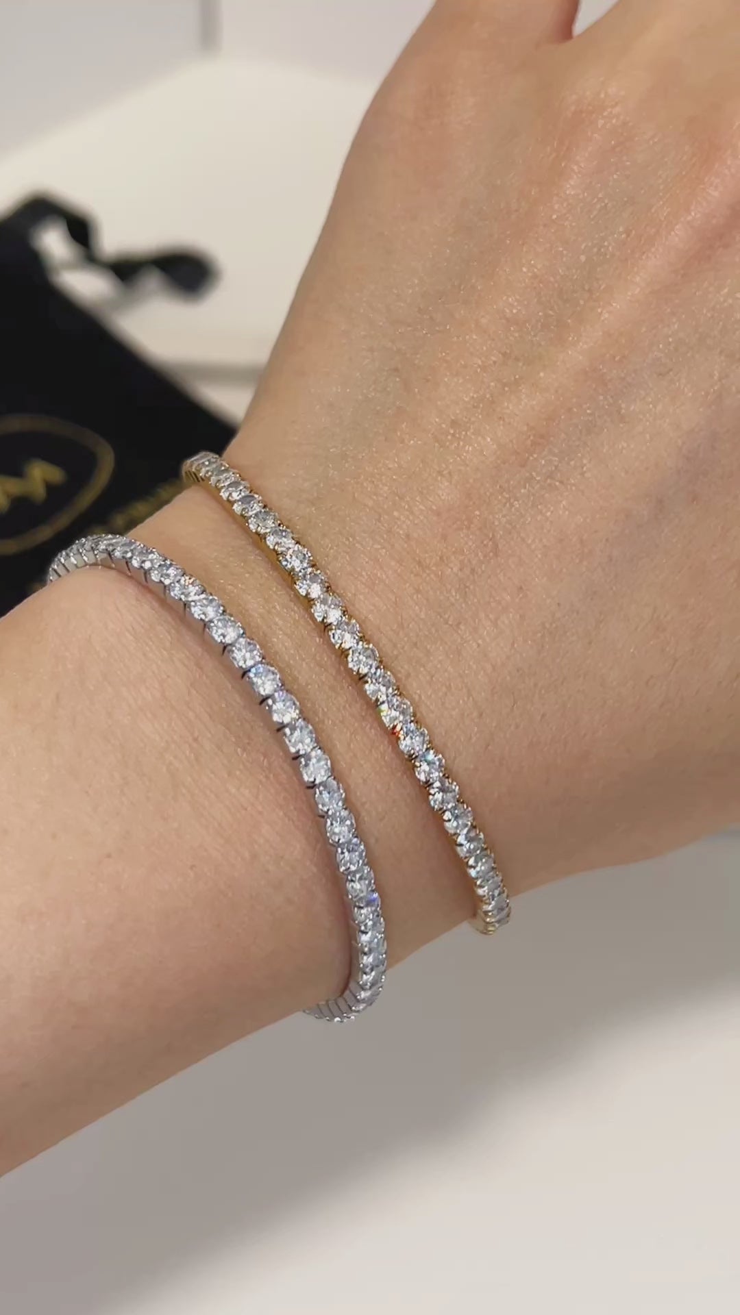A wrist wearing a silver and gold diamond tennis bracelet with a black pouch in the background
