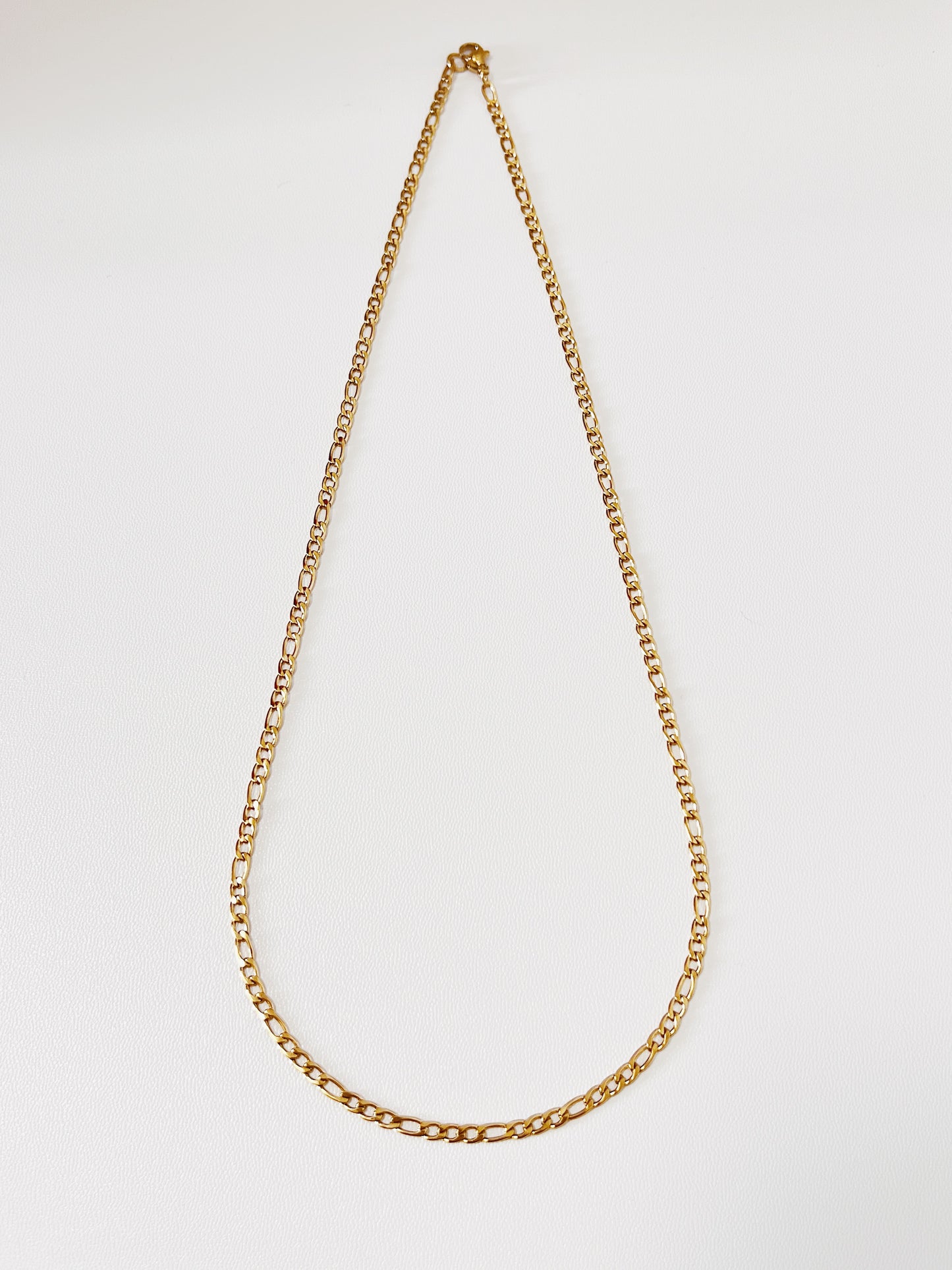 Top down shot of the Rhea Figaro chain links, accentuating the deep saturation of the 18k gold PVD finish under soft studio lighting.