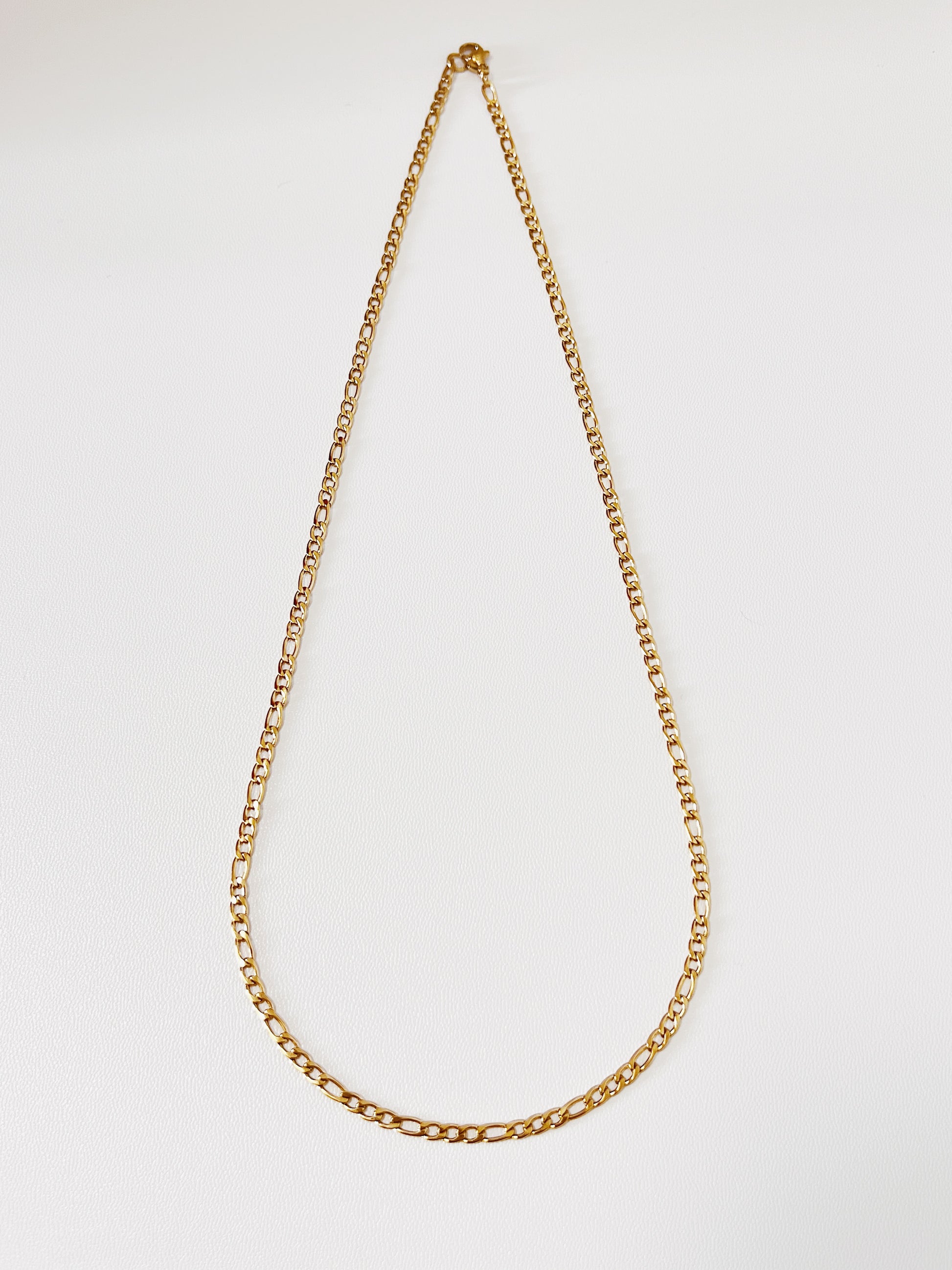 Top down shot of the Rhea Figaro chain links, accentuating the deep saturation of the 18k gold PVD finish under soft studio lighting.