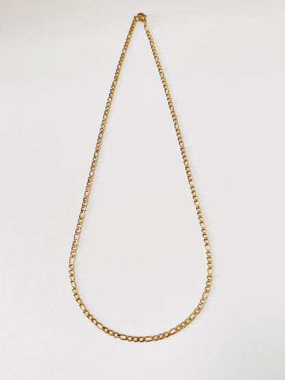 Top down shot of the Rhea Figaro chain links, accentuating the deep saturation of the 18k gold PVD finish under soft studio lighting.