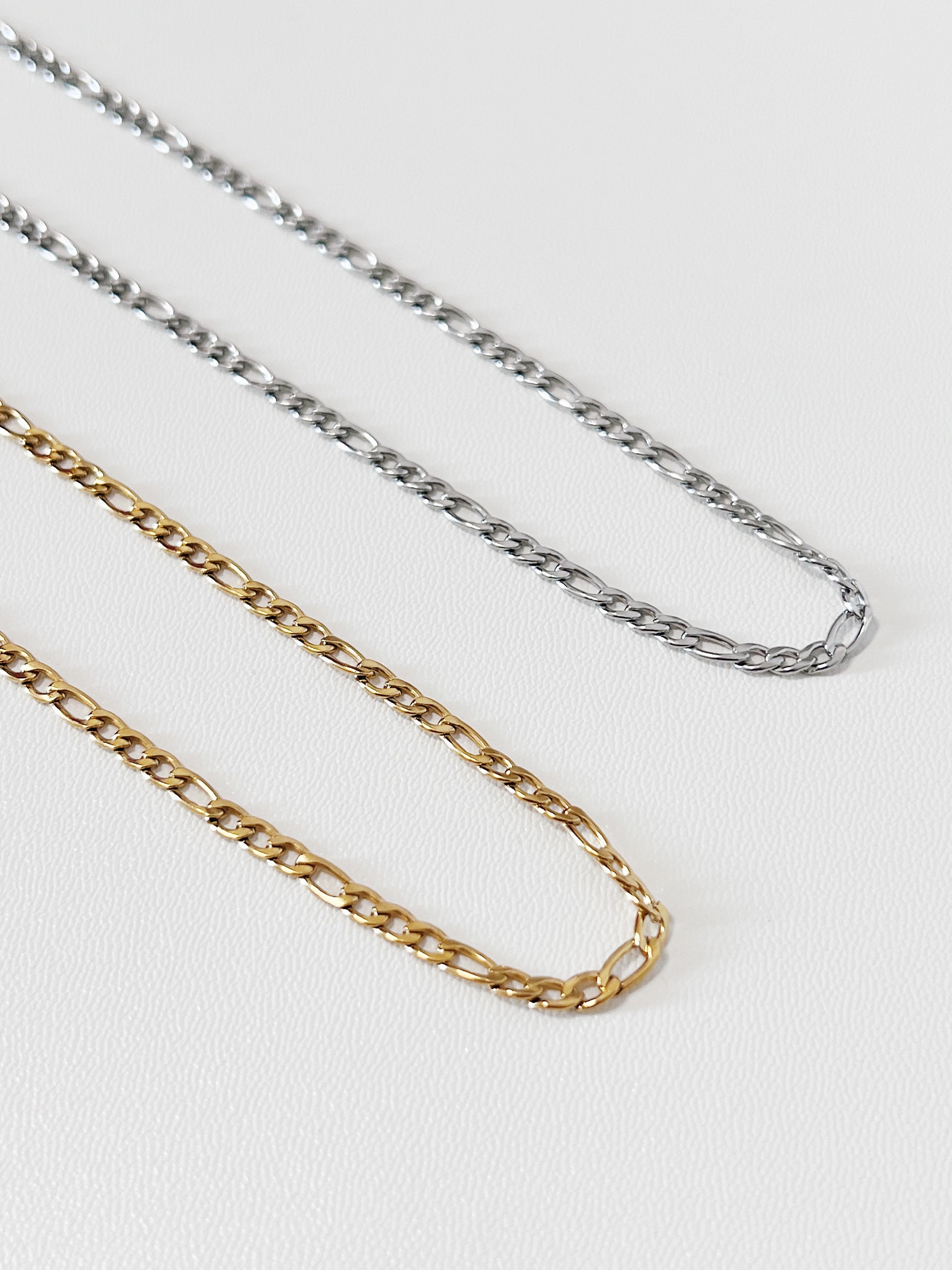 Overhead view of Rhea Figaro necklaces in both gold and silver laid side-by-side on white surface, displaying the rich contrast of the dual-metal collection.