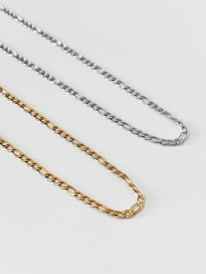Overhead view of Rhea Figaro necklaces in both gold and silver laid side-by-side on white surface, displaying the rich contrast of the dual-metal collection.