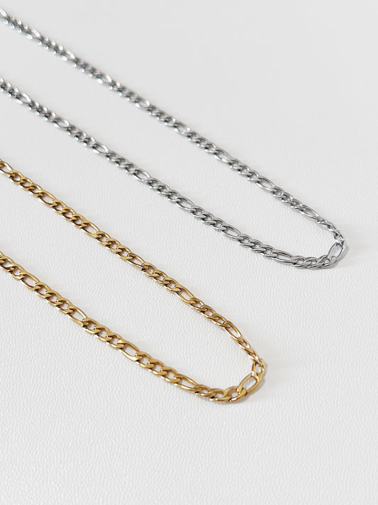 Overhead view of Rhea Figaro necklaces in both gold and silver laid side-by-side on white surface, displaying the rich contrast of the dual-metal collection.