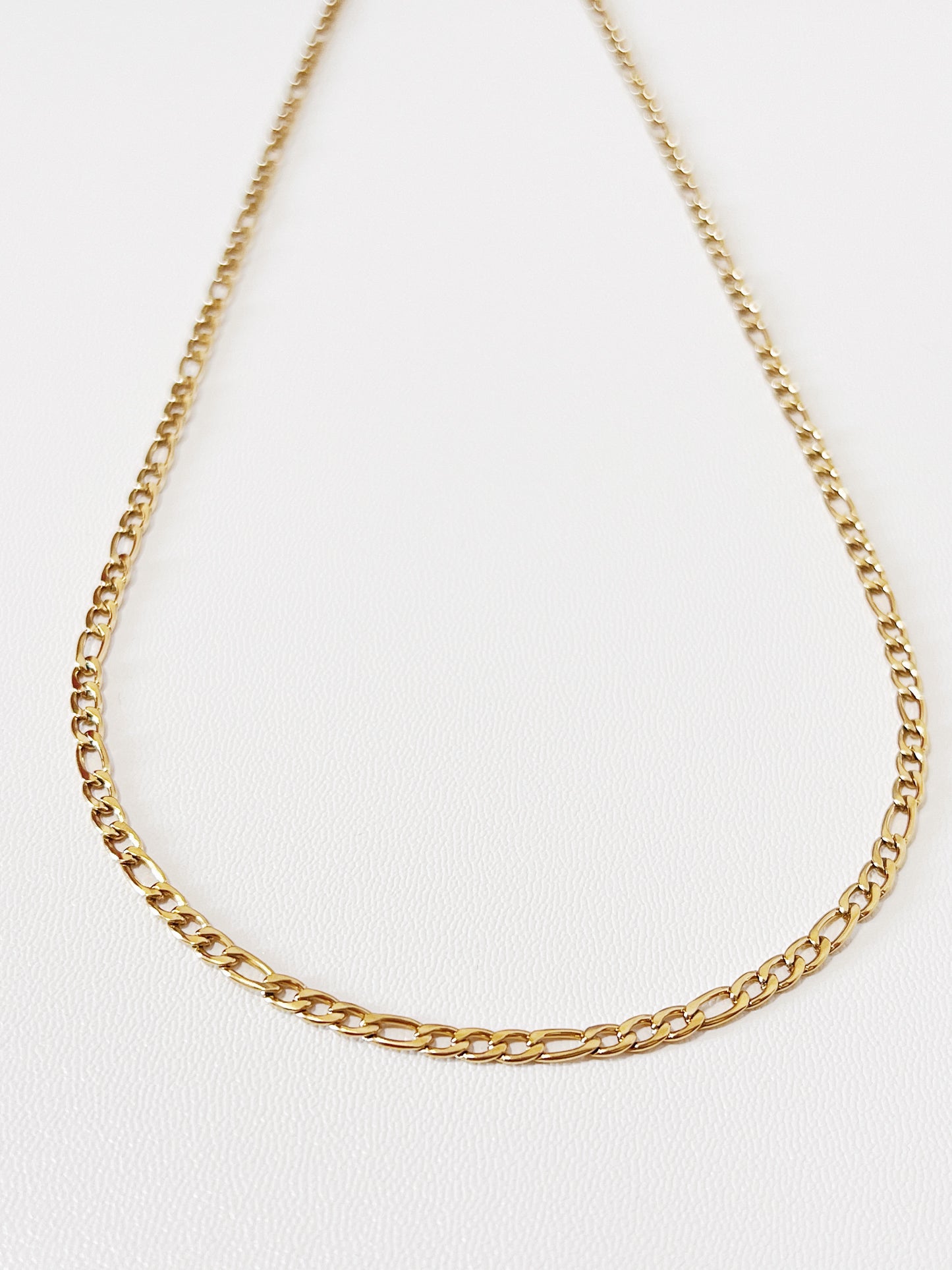 Close-up product photography of Rhea gold Figaro chain necklace resting in a neat circular layout on a neutral surface, showcasing the 3:1 alternating link pattern.