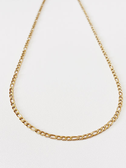 Close-up product photography of Rhea gold Figaro chain necklace resting in a neat circular layout on a neutral surface, showcasing the 3:1 alternating link pattern.