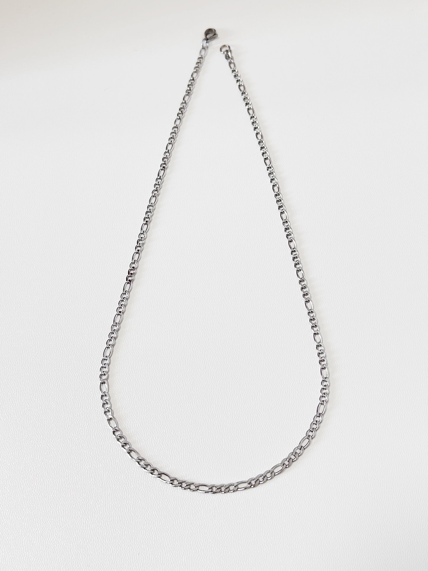 Studio detail shot of Rhea silver Figaro link chain, highlighting the high-polish mirror finish and the crisp edges of the flat stainless steel links.