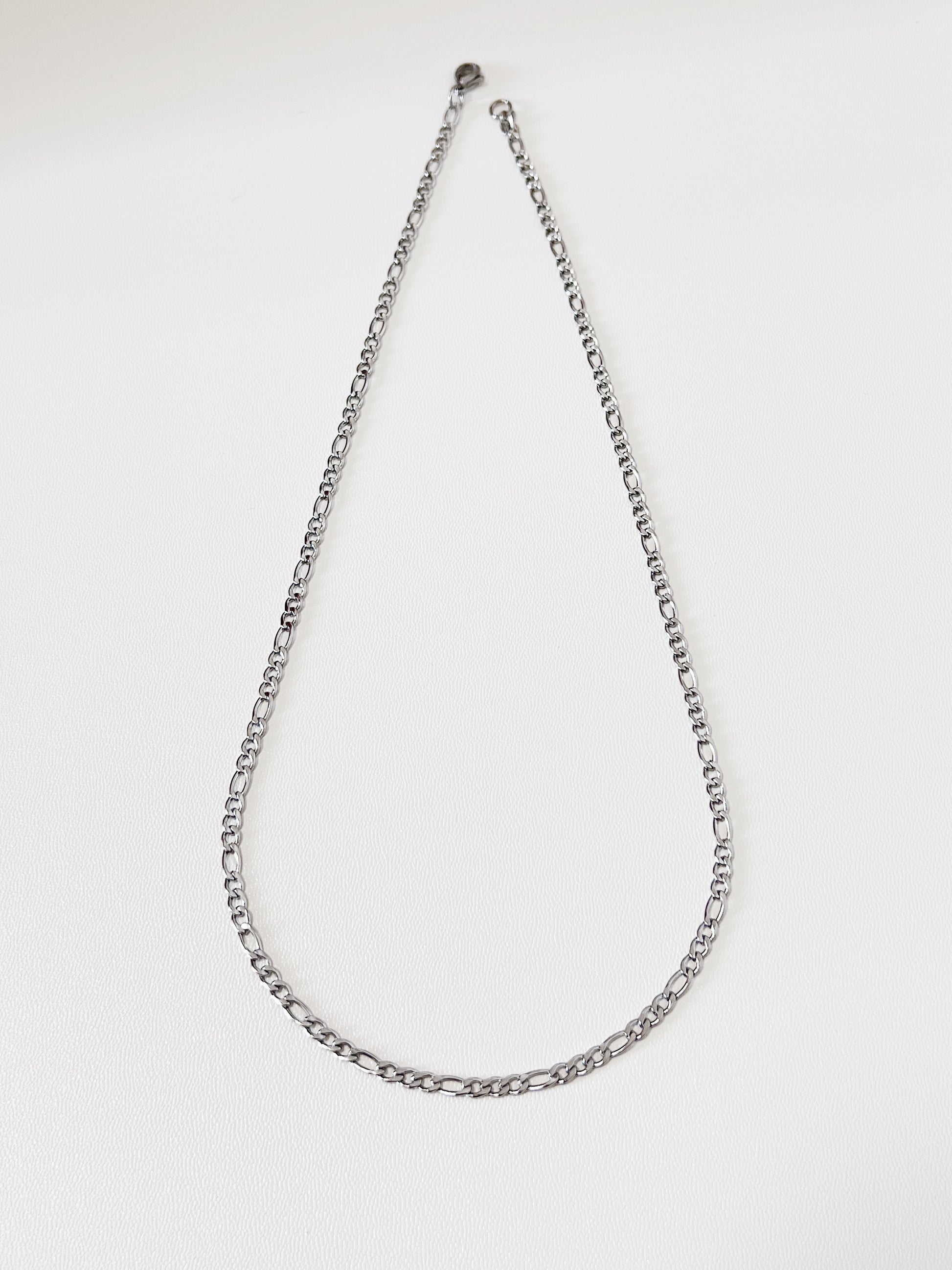 Studio detail shot of Rhea silver Figaro link chain, highlighting the high-polish mirror finish and the crisp edges of the flat stainless steel links.