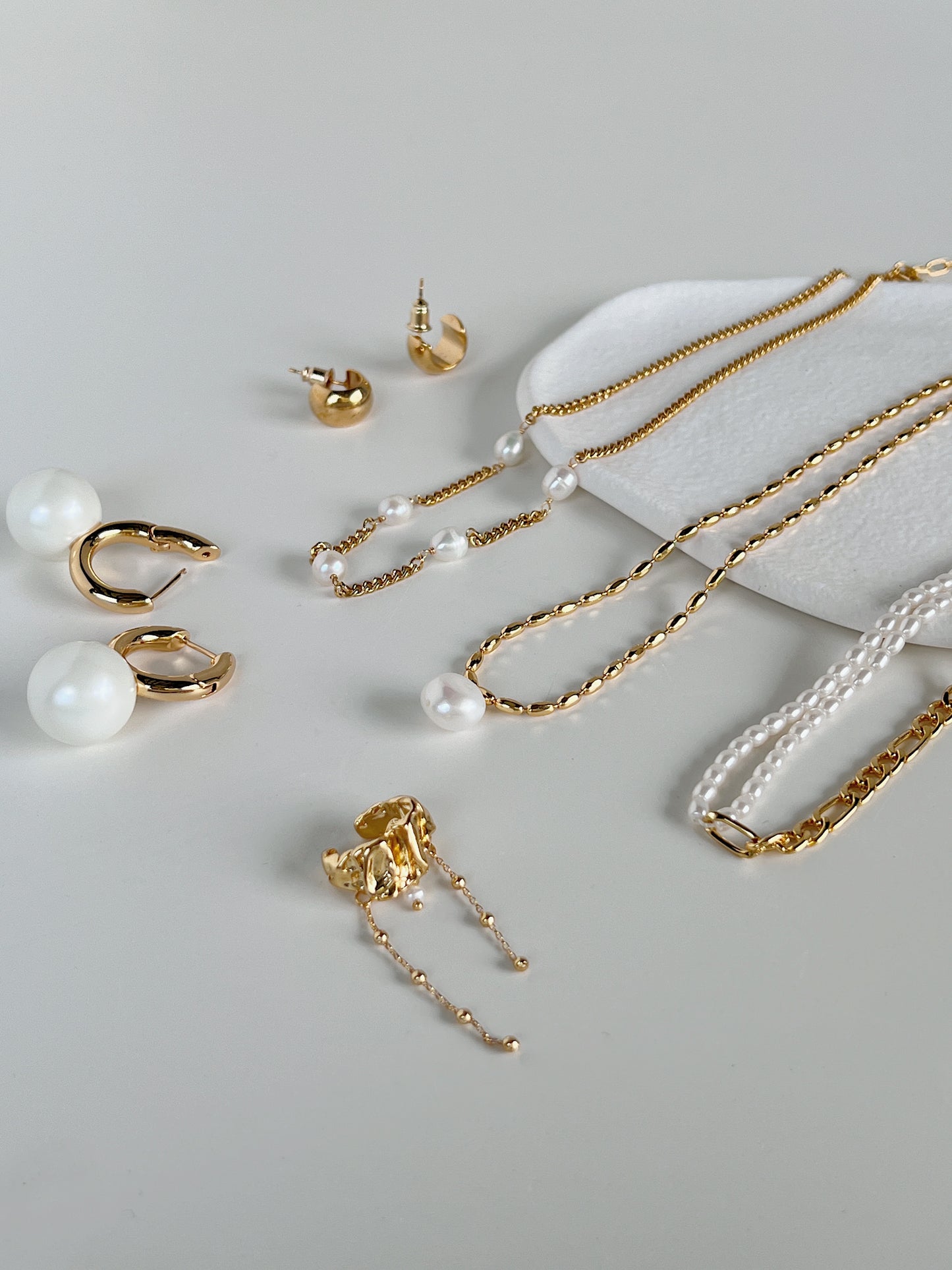 A curated jewelry tray edit showcasing contemporary freshwater pearl drop necklaces, high shine tarnish resistant gold chains, textured statement ear cuffs, and organic spherical earrings for a complete lookbook capsule