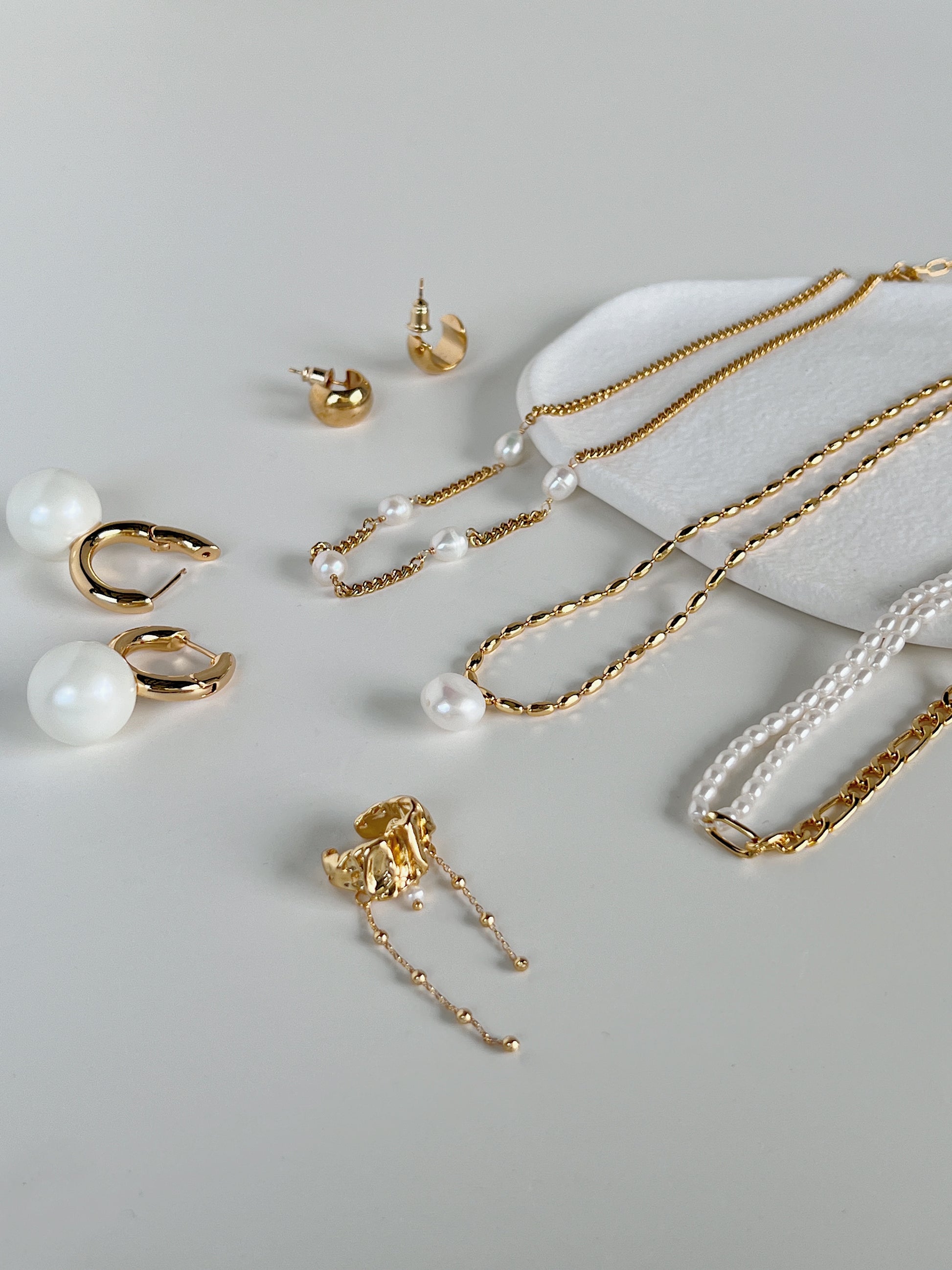 A curated jewelry tray edit showcasing contemporary freshwater pearl drop necklaces, high shine tarnish resistant gold chains, textured statement ear cuffs, and organic spherical earrings for a complete lookbook capsule