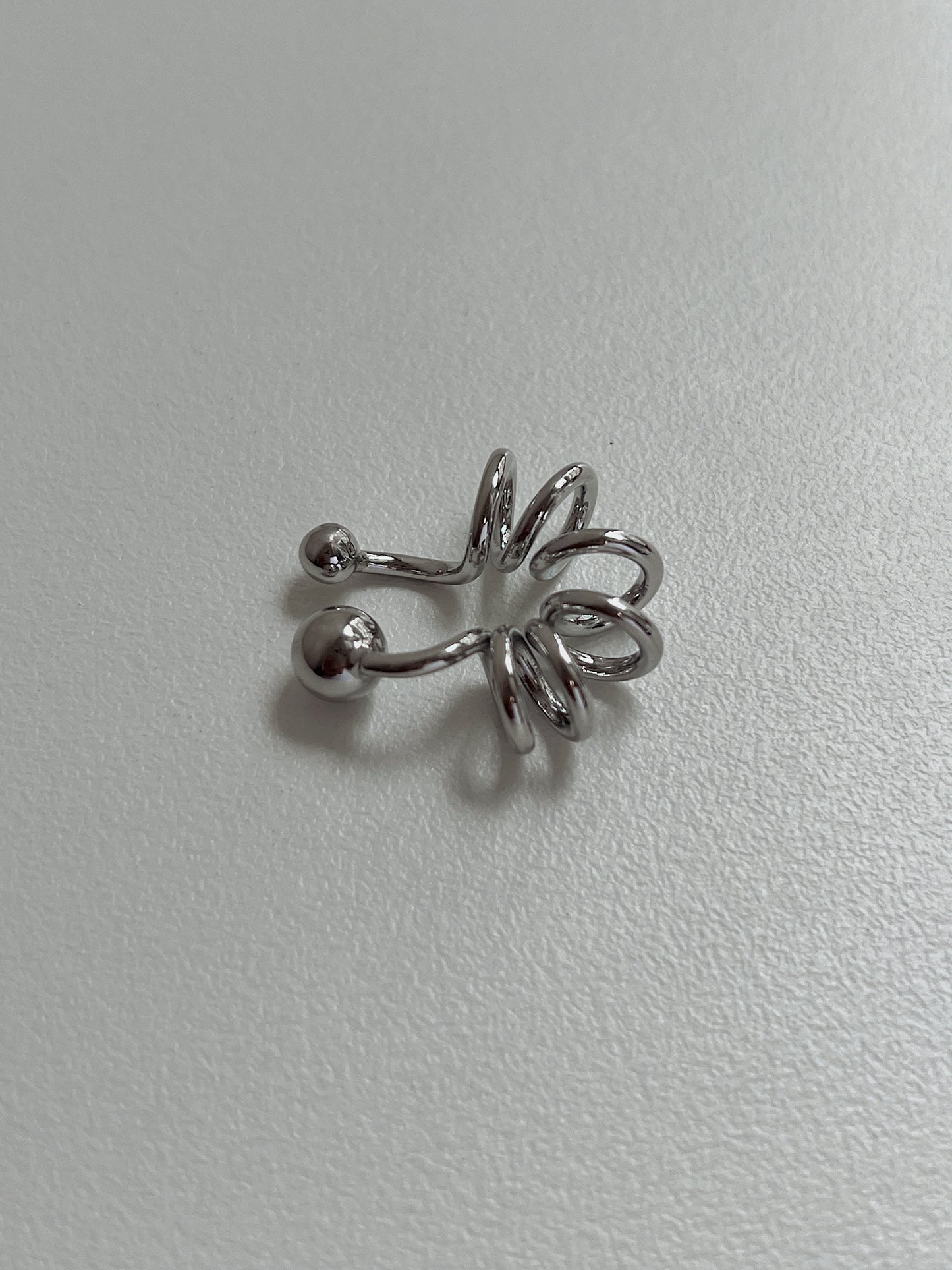 Silver Lis Swirl Ear Cuff on a minimalist, light-grey textured background.