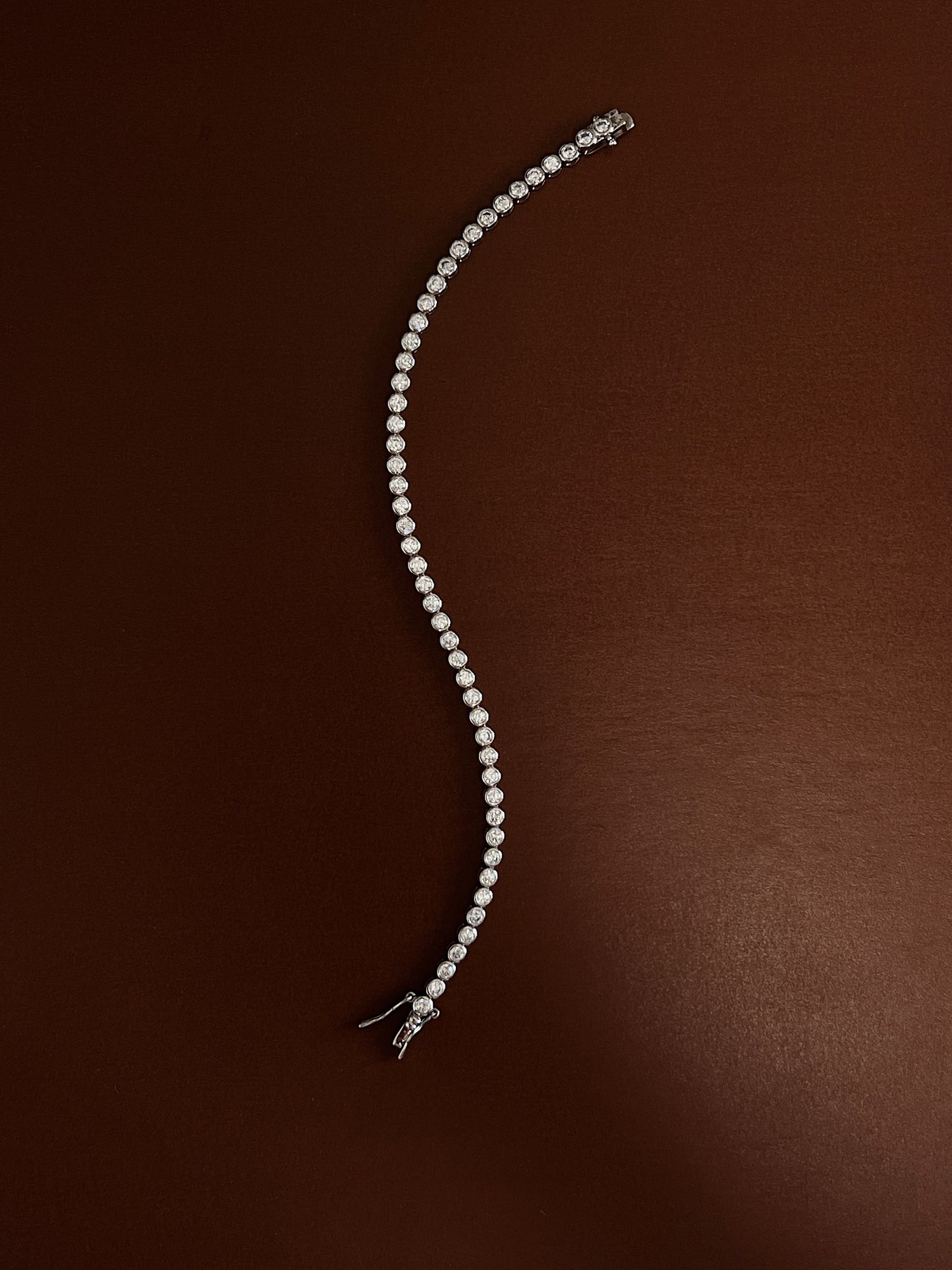 Rhodium-plated bezel tennis bracelet laid in a fluid serpentine curve on a rich espresso backdrop showcasing its flexible link drape.