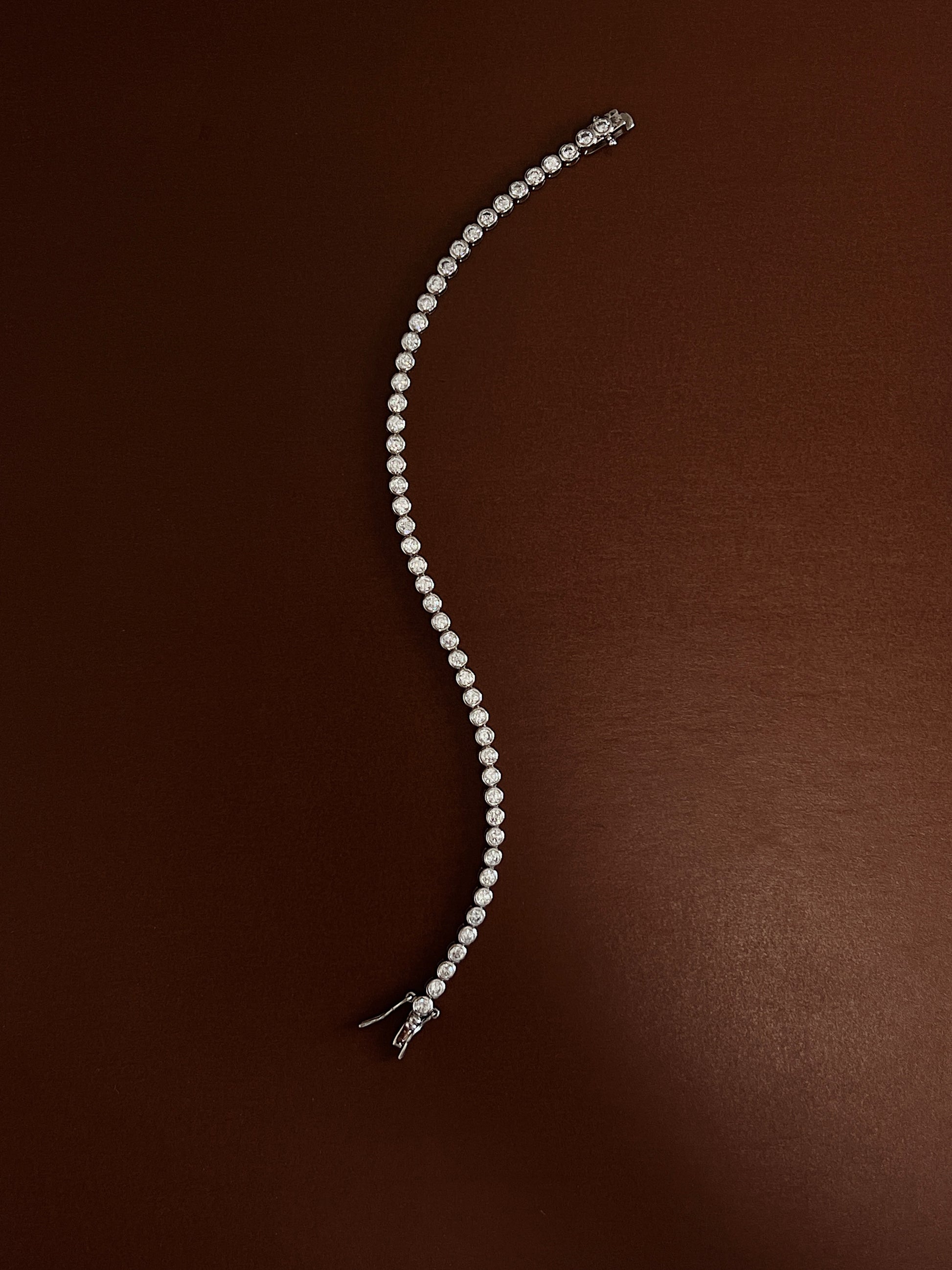 Rhodium-plated bezel tennis bracelet laid in a fluid serpentine curve on a rich espresso backdrop showcasing its flexible link drape.