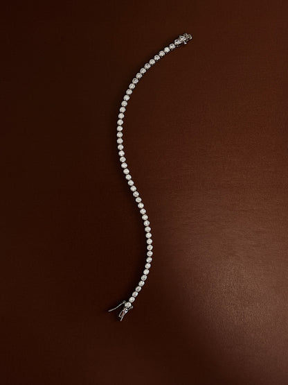 Rhodium-plated bezel tennis bracelet laid in a fluid serpentine curve on a rich espresso backdrop showcasing its flexible link drape.