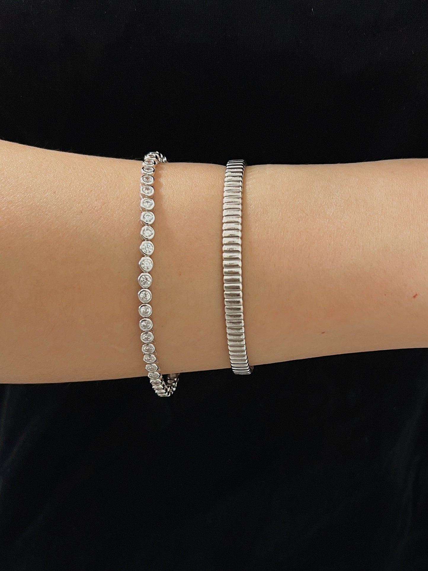 Horizontal wrist view showing the smooth, flush bezel-set stones of a silver tennis bracelet paired with a textured metallic cuff.