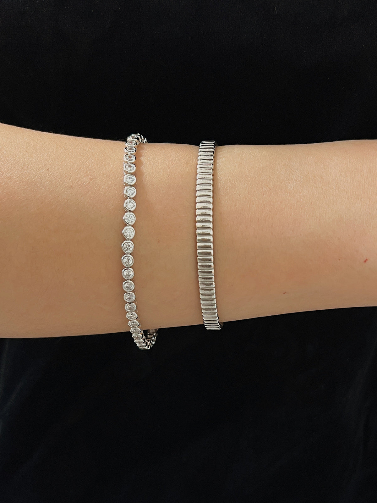 Horizontal wrist view showing the smooth, flush bezel-set stones of a silver tennis bracelet paired with a textured metallic cuff.