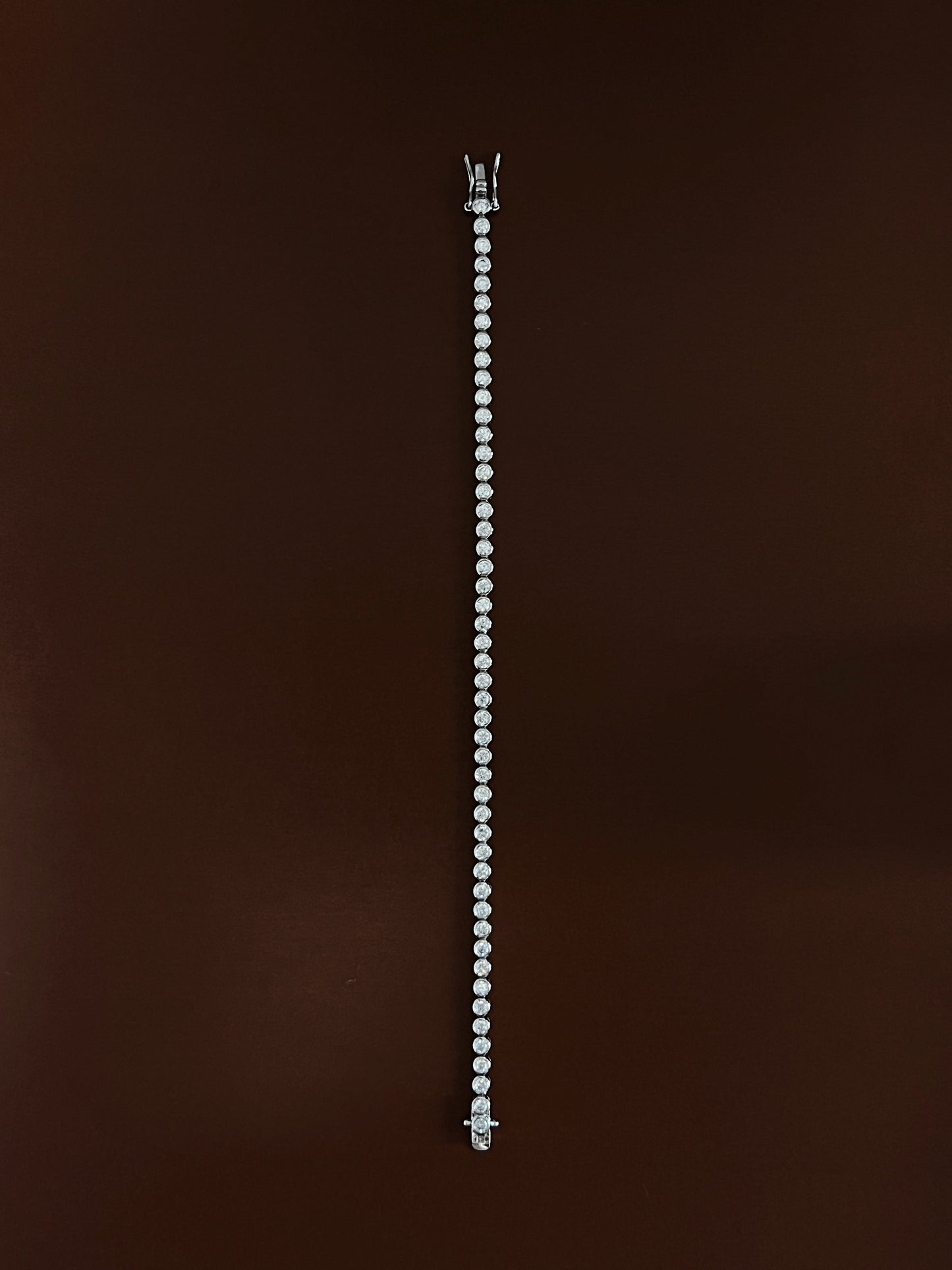 High-luster rhodium finish gleaming across the round metallic bezels of a silver tennis bracelet laid straight on dark wood color surface.