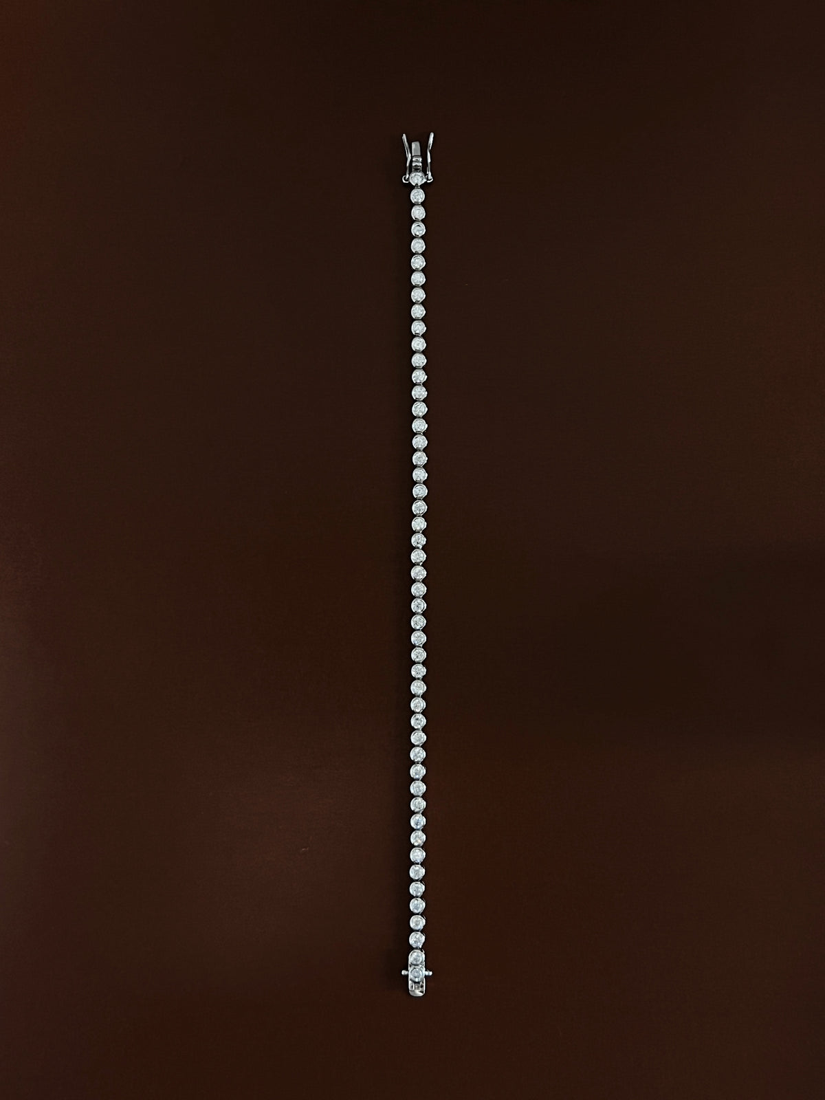 High-luster rhodium finish gleaming across the round metallic bezels of a silver tennis bracelet laid straight on dark wood color surface.