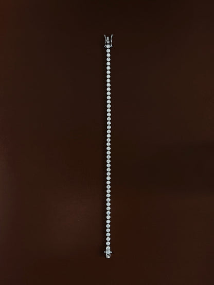 High-luster rhodium finish gleaming across the round metallic bezels of a silver tennis bracelet laid straight on dark wood color surface.