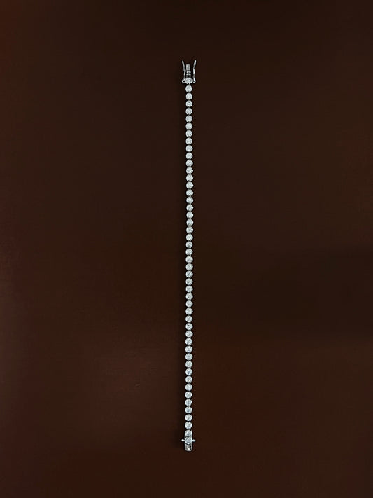 High-luster rhodium finish gleaming across the round metallic bezels of a silver tennis bracelet laid straight on dark wood color surface.