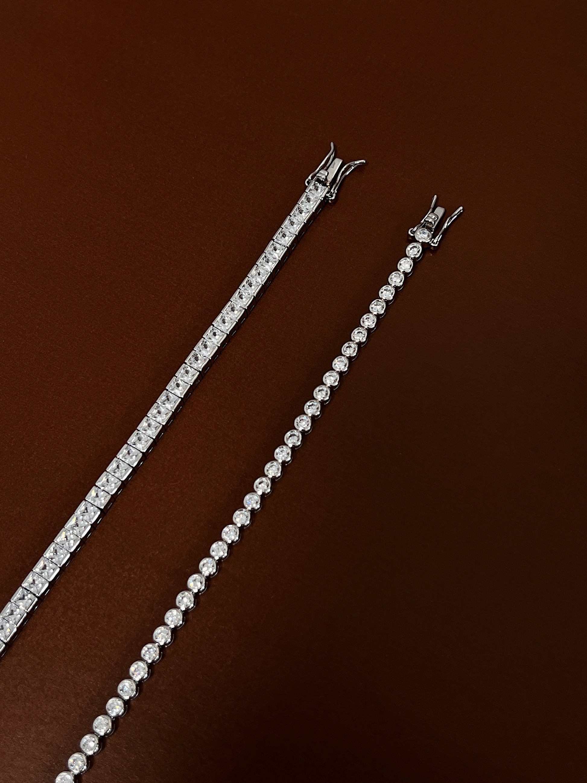 Side-by-side detail of a square-cut tennis bracelet and a round bezel-set tennis bracelet on a dark brown surface showing box clasp details.