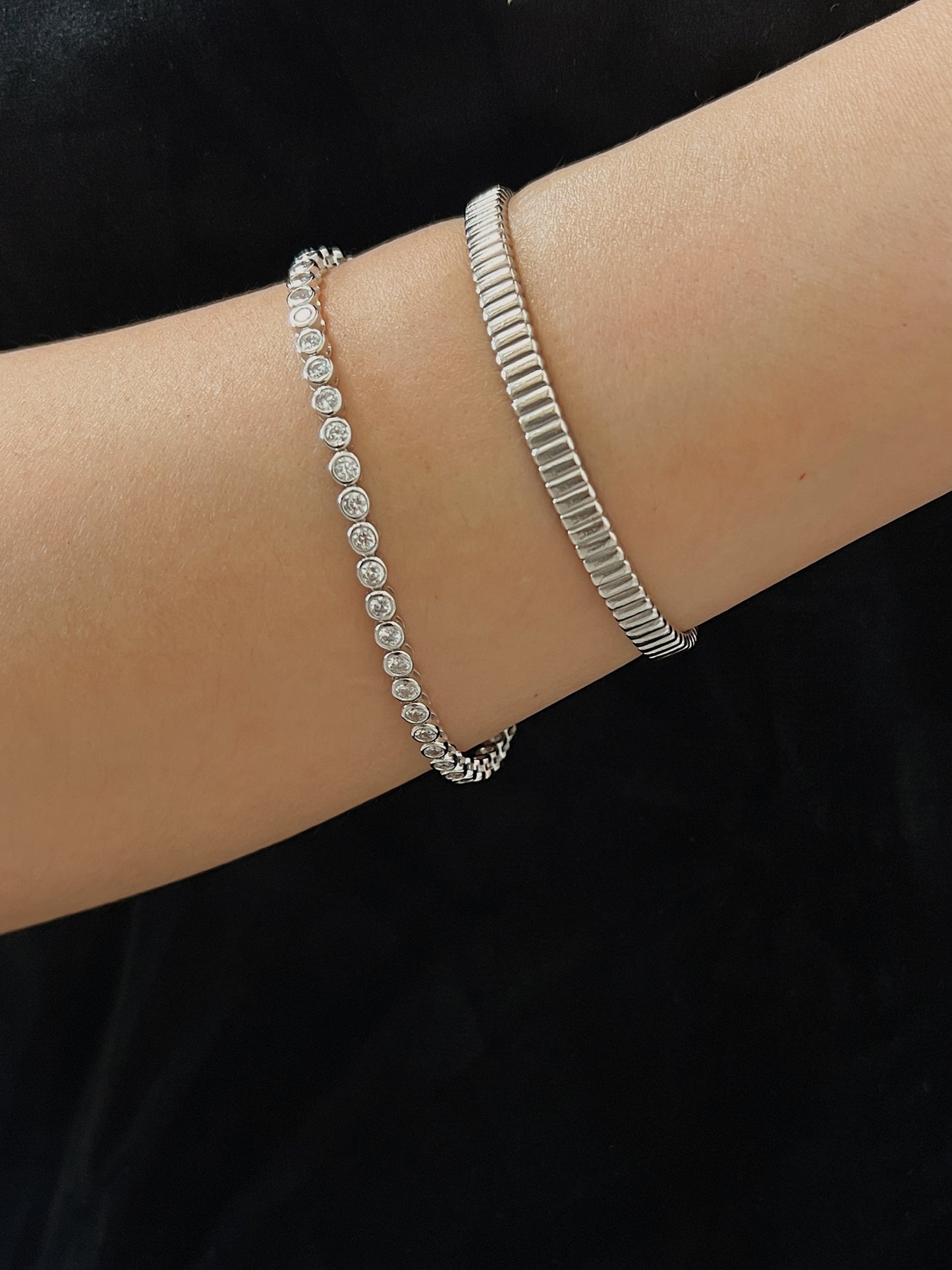 Close-up shot of a silver round bezel tennis bracelet stacked alongside a textured vede ribbed metal cuff on a model's wrist.