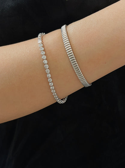 Close-up shot of a silver round bezel tennis bracelet stacked alongside a textured vede ribbed metal cuff on a model's wrist.