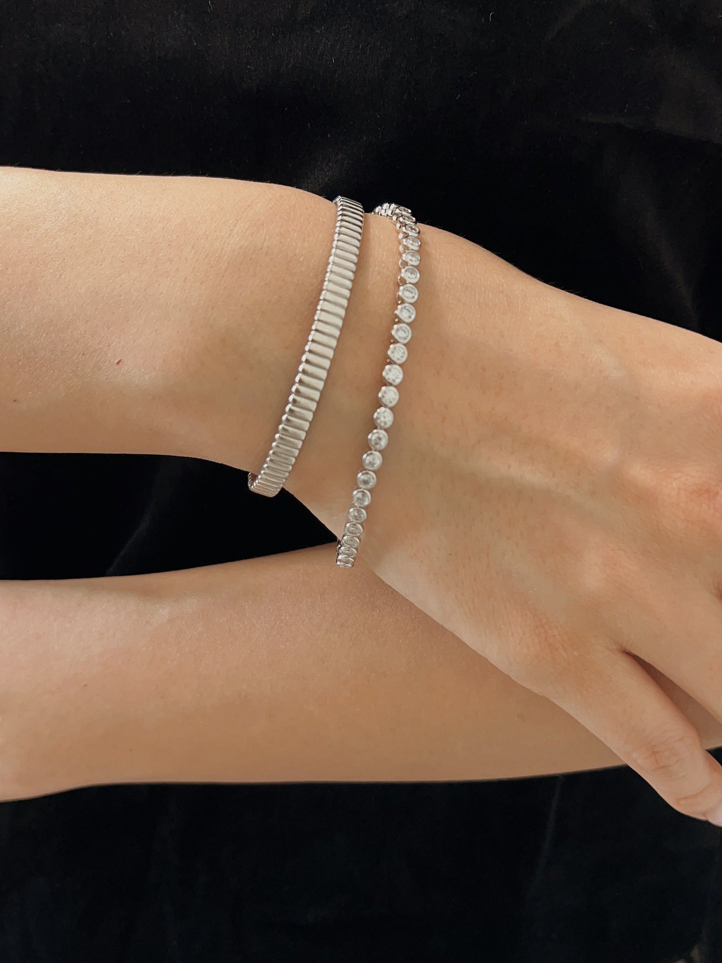 Model wearing a round bezel tennis bracelet stacked with a silver ridged cuff against a black velvet outfit.