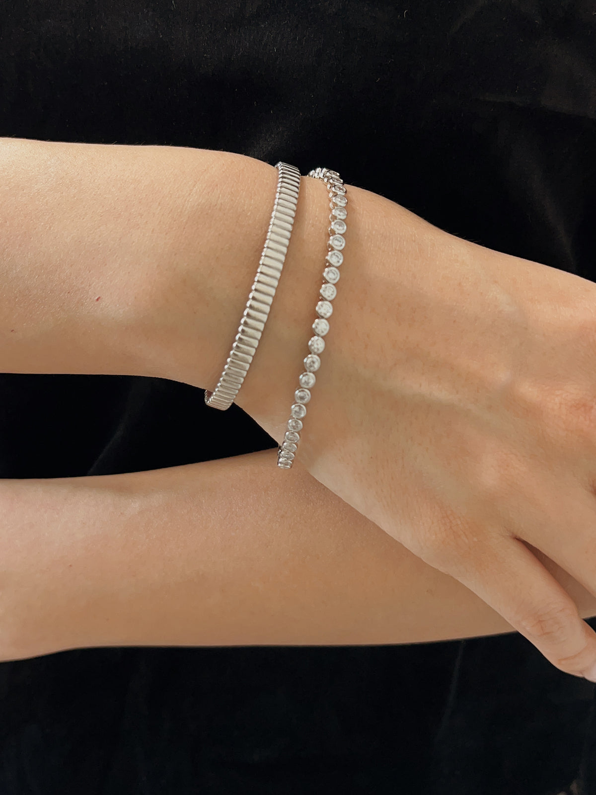 Model wearing a round bezel tennis bracelet stacked with a silver ridged cuff against a black velvet outfit.