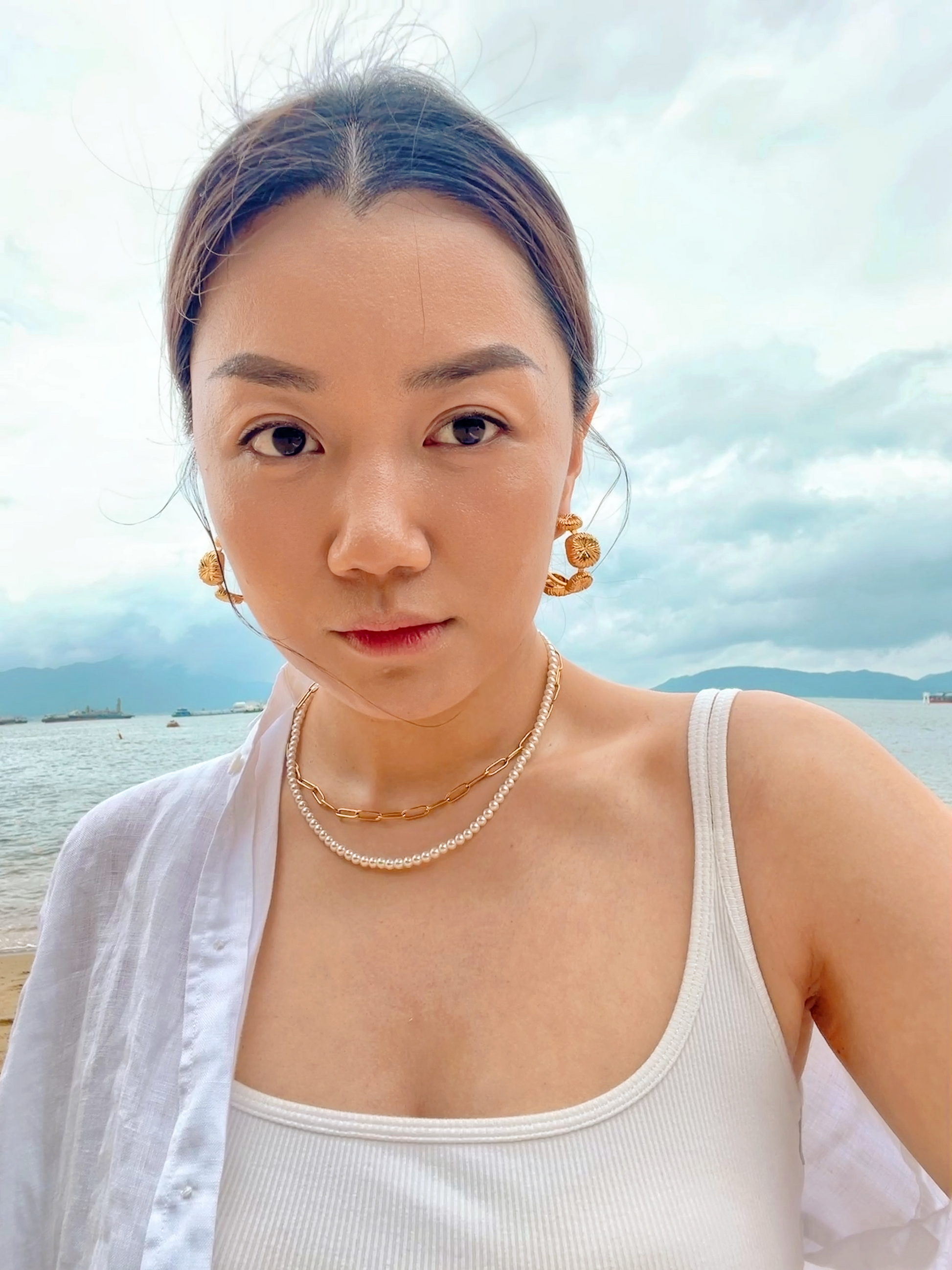 Close up of woman styling the marianna tiny pearl choker necklace with a relaxed white linen shirt on a sandy beach