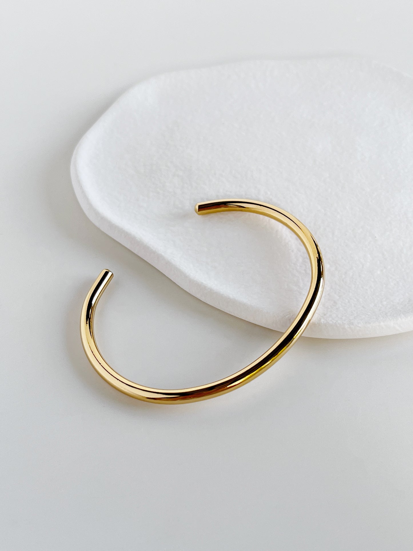 Overhead studio photography of the Ines tarnish-resistant gold bangle on a modern white background, demonstrating the sleek curve and uniform thickness.