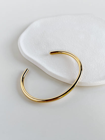Overhead studio photography of the Ines tarnish-resistant gold bangle on a modern white background, demonstrating the sleek curve and uniform thickness.
