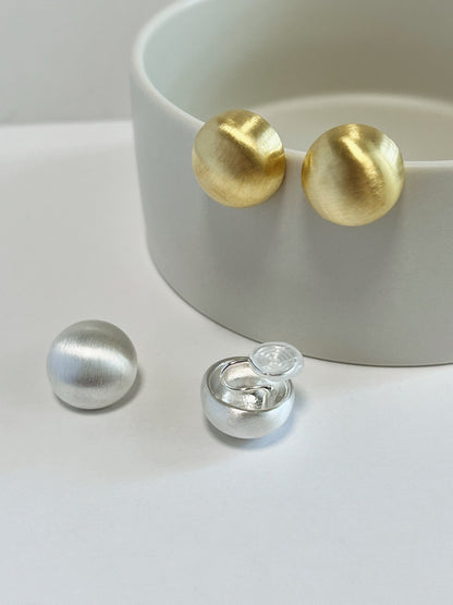 Discreet Japanese coil wire clip with clear silicone comfort pad revealed alongside gold and silver brushed dome earrings on a ceramic dish.