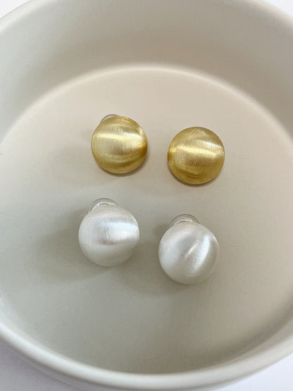 Overhead view comparing 16K gold plated and cool rhodium silver Tyla 18mm non-pierced dome earrings arranged inside an off-white tray.