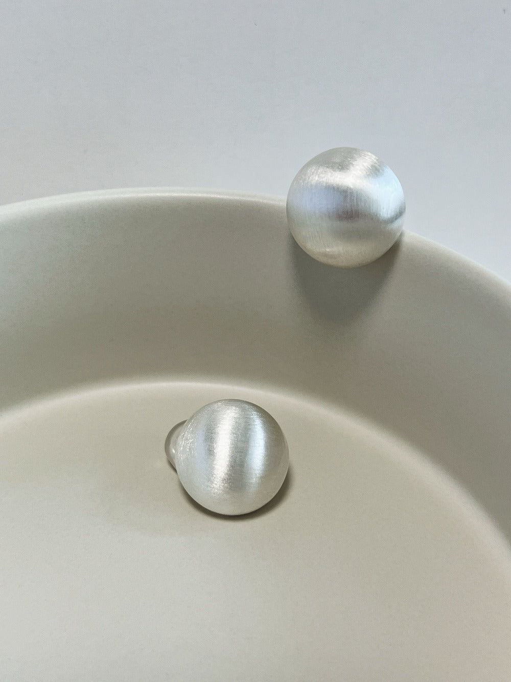 Soft studio light diffusing across the linear brushed micro-texture of rhodium-plated silver dome clip-on earrings resting on ceramic stoneware.