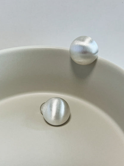 Soft studio light diffusing across the linear brushed micro-texture of rhodium-plated silver dome clip-on earrings resting on ceramic stoneware.