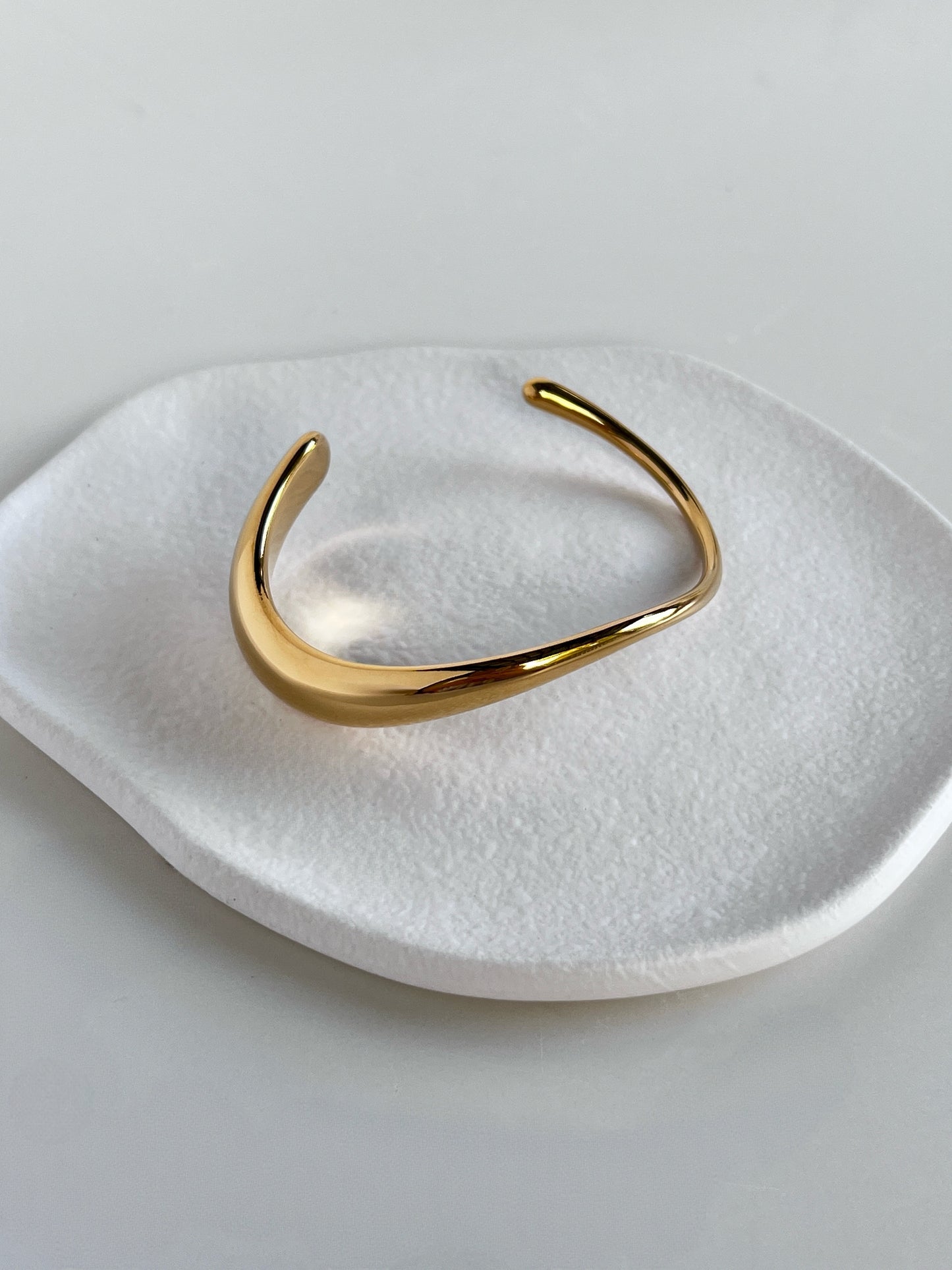A high-luster gold Vicky sculpted wave cuff bangle resting flat on a white textured ceramic dish, highlighting its fluid asymmetrical opening.