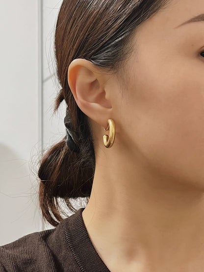 Three-quarter view of a model showcasing the secure, snug lobe fit of the Zora solid-body matte gold clip-on hoop earrings paired with a dark brown knit top.