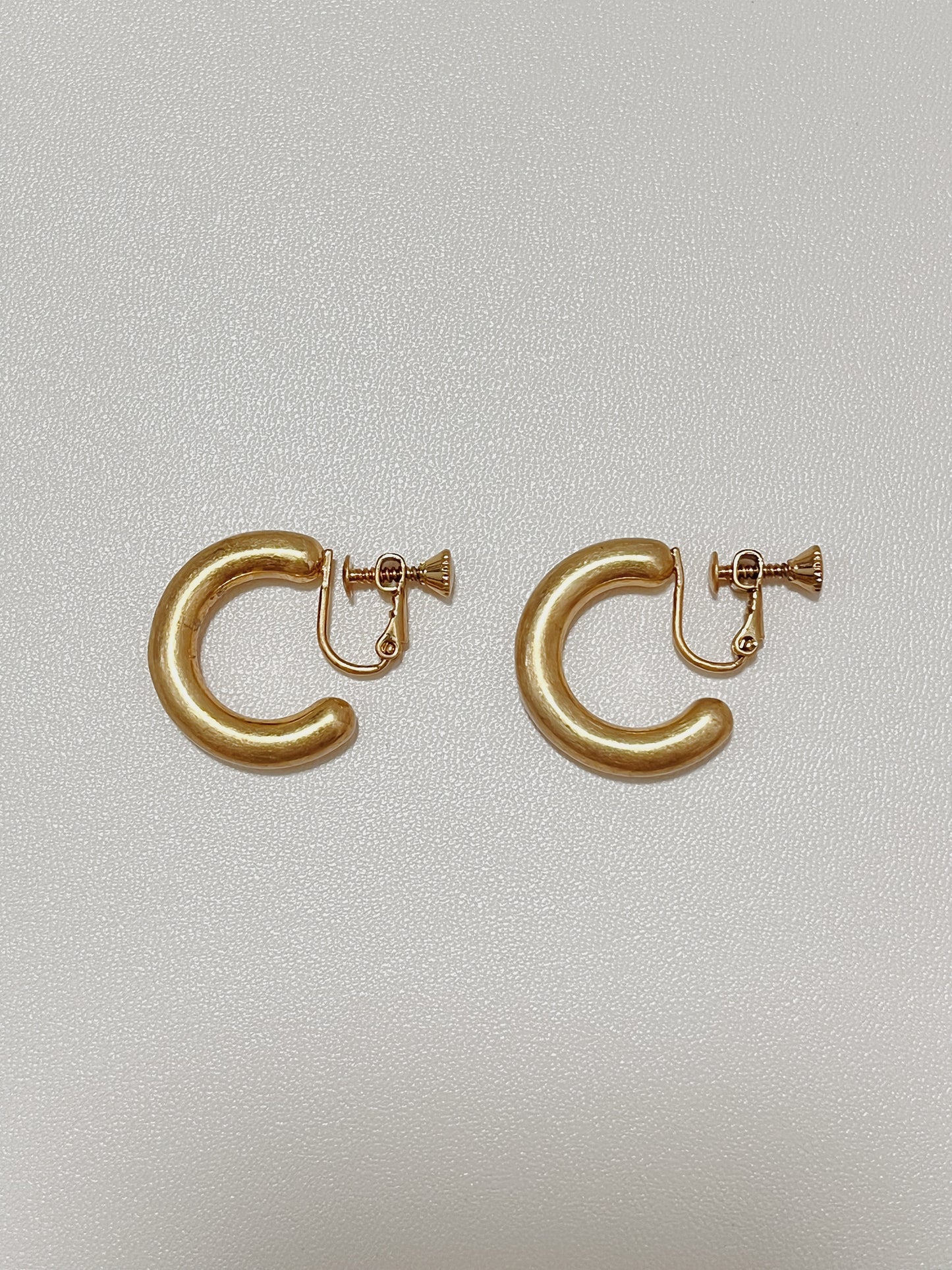 Side-by-side view of Zora solid-body brushed gold clip-on hoop earrings showing the custom matte finish and curved silhouette against a fine textured backdrop.